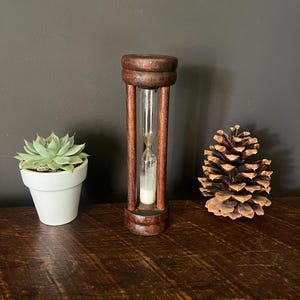 May include: A dark brown wooden hourglass with a clear glass center containing white sand, positioned between a small green succulent in a white ceramic pot and a large brown pinecone on a dark wooden surface.