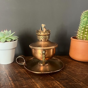 May include: A vintage copper oil lamp with a round base and a handle. The lamp has a small, decorative top with a wick. The lamp is sitting on a wooden surface, with a succulent and a cactus in the background.