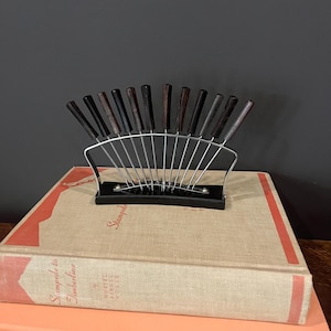 May include: A set of 12 silver skewers with dark wood handles, arranged in a fan shape on a black stand. The stand is sitting on top of two books.