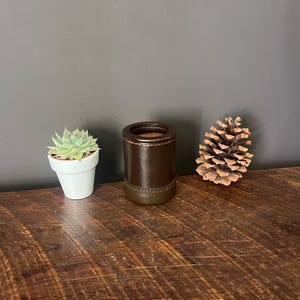 May include: A cylindrical dark brown leather-wrapped container with visible stitching detail, positioned between a small green succulent in a white ceramic pot and a natural brown pinecone on a dark wooden surface.