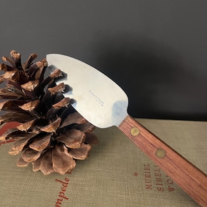 May include: A stainless steel cheese knife with a wooden handle, featuring brass rivets. The knife is from Hickory Farms. A large pinecone rests on a book with the word "Stampede" visible.