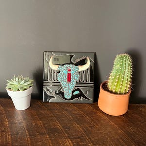May include: A square, decorative tile featuring a turquoise bull skull design with cream-colored horns and a red accent. The tile is set against a dark gray background. A small succulent and cactus in pots are on either side.