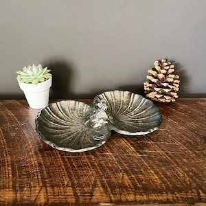 May include: A silver-toned, double-dish decorative bowl with a seashell design. The bowl is on a wooden surface, with a small potted succulent and a pine cone in the background.