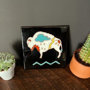 May include: Square black tile featuring a white bison with colorful accents. The bison has a cloud and lightning bolt design. A turquoise zig-zag line is at the bottom. The tile is on a wooden surface.