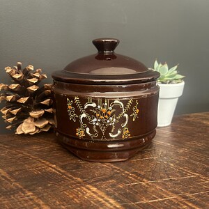 May include: A brown ceramic jar with a lid, decorated with a gold floral design. The jar has a glossy finish and is sitting on a wooden surface.