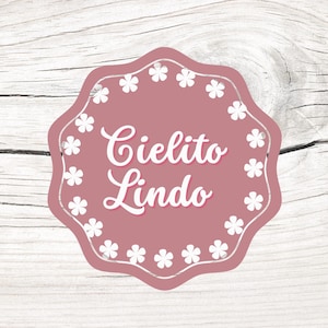 Könnte beinhalten: Ein rundes, rosa und weißes Etikett mit einem gewellten Rand und weißen Blumen um den Rand. Der Text "Cielito Lindo" ist in weißer Kursivschrift geschrieben.