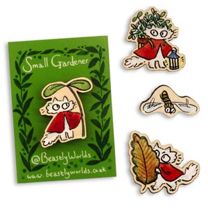 Pode incluir: Pins de madeira com um gato de desenho animado vestido com uma capa vermelha, com o tema de jardineiro. Um pin está em um cartão verde com o texto "Small Gardener". Os pins são pintados à mão com um design fantasioso.
