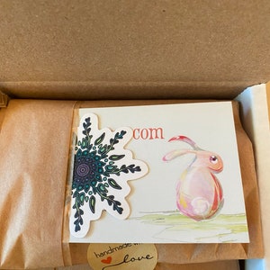 May include: A brown cardboard box with a brown paper package inside. The package has a sticker with a mandala design and a sticker that says "handmade with love" and a card with a watercolor illustration of a rabbit and the text "com".