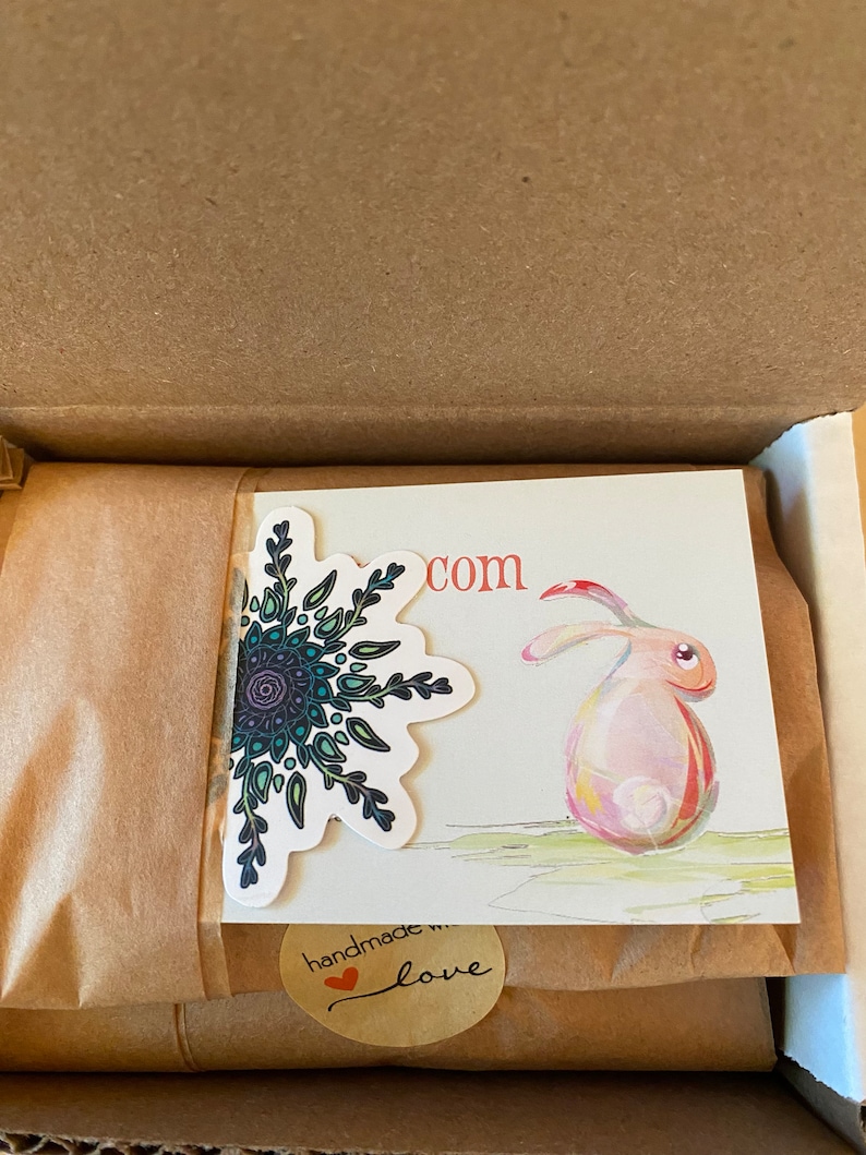 May include: A brown cardboard box with a brown paper package inside. The package has a sticker with a mandala design and a sticker that says "handmade with love" and a card with a watercolor illustration of a rabbit and the text "com".