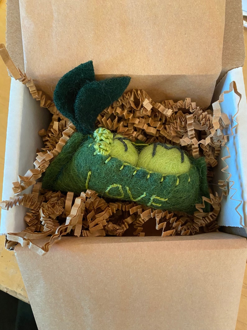 May include: A green felt pea pod with a stitched design that says "LOVE" inside a brown cardboard box with brown paper packing peanuts.