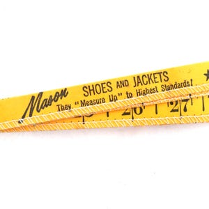 May include: Close-up of a vintage yellow measuring tape with black text that reads "Mason SHOES AND JACKETS". The text "They 'Measure Up' to Highest Standards!" is also visible. White stitching runs along the edges.