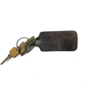May include: A set of keys on a metal ring with a dark brown leather key fob. The key fob is rectangular with rounded corners and appears to have some text or design on it. The keys are gold-coloured.