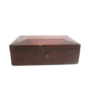 May include: A rectangular wooden box with a dark brown finish and a beveled lid featuring a lighter brown central panel. The front of the box displays a small, centered metal keyhole escutcheon and a visible key protruding from the lock mechanism.
