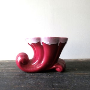 May include: A vintage, glossy, red ceramic planter in a horn shape. The planter has a wavy rim with a white and pink trim. The base has a curled design. The planter sits on a dark wooden surface.