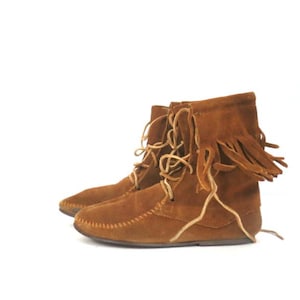 Puede incluir: Un par de botines de ante marrón con una construcción suave y flexible, que presenta una costura en la puntera estilo mocasín. Las botas incluyen un cierre frontal con cordones finos de color claro y un detalle de flecos decorativos.