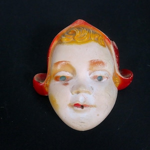 May include: A vintage wall-mounted mask featuring a pale face with painted blue eyes, rosy cheeks, and red lips. The figure wears a bright red hood with yellow hair visible at the forehead. The mask has a smooth, molded surface with visible wear.
