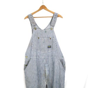 May include: Blue and white hickory striped denim overalls featuring a bib pocket with a rectangular OshKosh B'gosh brand label, metal buttons at the sides and shoulder straps, and a front fly opening, all displayed on a wooden hanger.