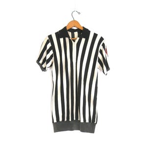 May include: A black and white striped short-sleeve shirt with a black collar and a partial zipper. The shirt has a small red emblem on the sleeve and a gray bottom hem. The shirt is hanging on a wooden hanger.