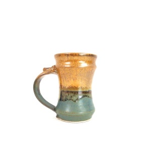 May include: A ceramic mug featuring a two-tone glaze with a speckled golden-brown upper section and a solid muted teal lower section. The mug has a curved handle and a slightly flared rim, showcasing a smooth, glossy finish throughout the exterior.