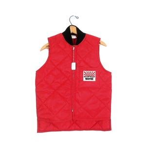 May include: A red quilted vest with a black collar and a full-length zipper. The vest features a small square patch on the chest with the word "WAYNE" and a rising sun design. The vest is hanging on a wooden hanger.