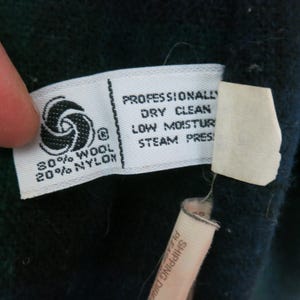 Puede incluir: Primer plano de una etiqueta de tela blanca con texto negro, que incluye "80% LANA 20% NYLON" y "LIMPIEZA EN SECO PROFESIONAL". La etiqueta tambi&eacute;n presenta un s&iacute;mbolo de lana. La etiqueta est&aacute; adherida a una tela azul oscuro.
