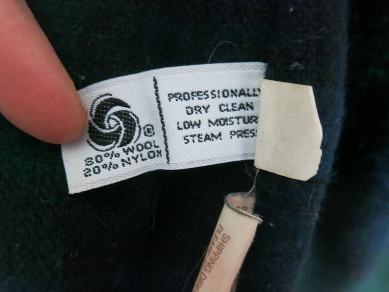 Puede incluir: Primer plano de una etiqueta de tela blanca con texto negro, que incluye "80% LANA 20% NYLON" y "LIMPIEZA EN SECO PROFESIONAL". La etiqueta tambi&eacute;n presenta un s&iacute;mbolo de lana. La etiqueta est&aacute; adherida a una tela azul oscuro.