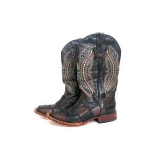 May include: A pair of black and brown leather cowboy boots featuring a patchwork design on the foot and intricate decorative stitching in light tan and black thread on the tall shaft, finished with a low stacked heel and a rounded toe profile.