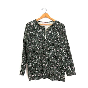 May include: A dark green long-sleeved shirt with a floral design. The shirt features a Henley neckline with three buttons. The floral pattern includes white and pink flowers and green leaves. The shirt is suspended from a wooden hanger.