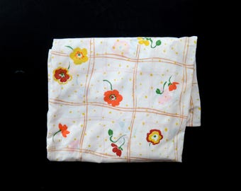 Puede incluir: Tela blanca doblada con un estampado floral. El diseño presenta flores naranjas, amarillas y rojas con tallos verdes, sobre una cuadrícula de líneas naranjas y puntos amarillos. La tela está doblada, mostrando su textura.