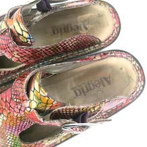 May include: Close-up of a pair of Alegria brand clogs. The clogs feature a colorful snakeskin pattern in shades of red, pink, yellow, and green. The interior of the shoe is a neutral beige color with the brand name printed on the insole.