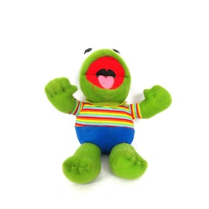 May include: A plush toy frog with a wide open mouth, green body, and blue shorts. The frog has a rainbow striped shirt and green arms and legs. The frog is sitting with its arms raised.