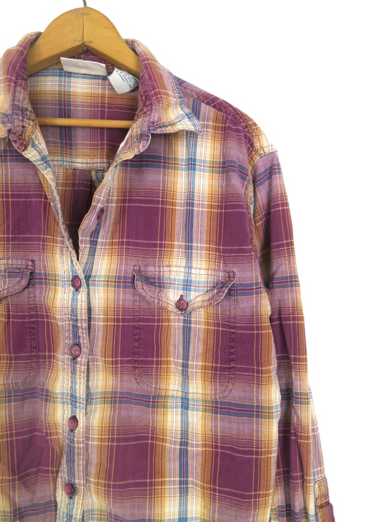 May include: A purple, yellow, and blue plaid button-down shirt with two chest pockets. The shirt is hanging on a wooden hanger against a white background. The plaid pattern is a mix of colors.