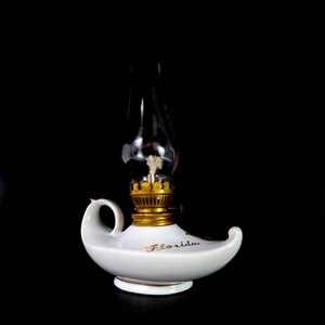 May include: A white ceramic oil lamp with a clear glass chimney and a gold-colored metal base. The word "Florida" is written in gold script on the lamp's body. The lamp has a handle and a curved spout.