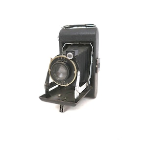 May include: A vintage black folding camera with a bellows mechanism, a circular lens housing labeled Kodak Anastigmat, and a metallic drop-down front panel. The camera body features a textured black finish, a top handle, and the word Kodak printed vertically.