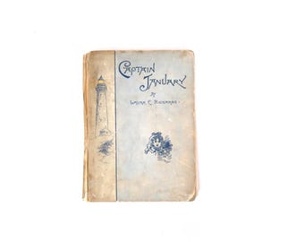 Puede incluir: Un libro de tapa dura antiguo titulado Captain January de Laura E. Richards. La cubierta es de color azul claro y tostado, con una ilustración azul oscuro de un faro a la izquierda y un pequeño retrato de una persona en el centro con texto azul.
