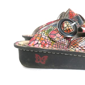May include: Close-up of a colorful sandal with a reptile-like pattern in shades of pink, orange, and green. The sandal has a black sole with a red butterfly emblem and a silver buckle.