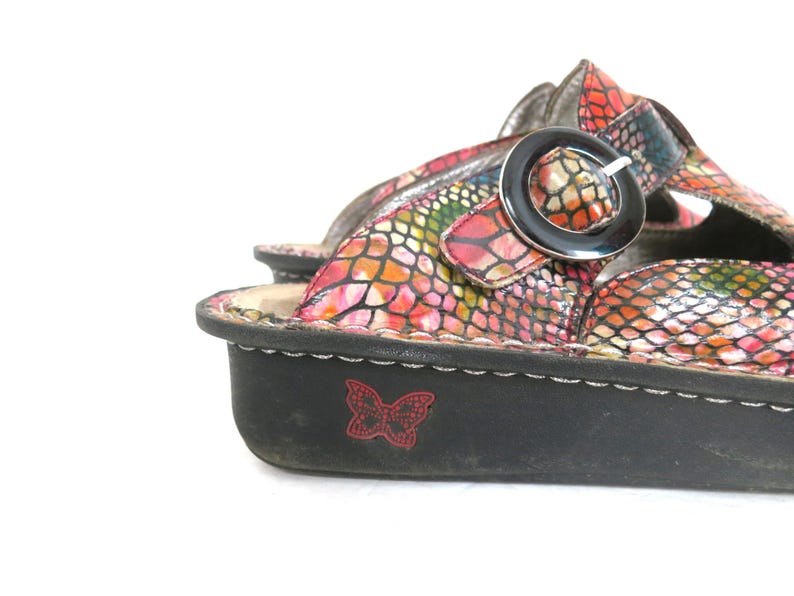 May include: Close-up of a colorful sandal with a reptile-like pattern in shades of pink, orange, and green. The sandal has a black sole with a red butterfly emblem and a silver buckle.