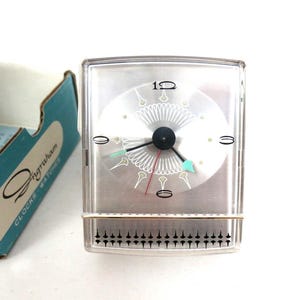 May include: A rectangular Ingraham alarm clock with a clear plastic casing, a silver face featuring black numerals at 12, 3, 6, and 9, and a decorative white pattern. It includes black hour and minute hands, a thin red second hand, and a light blue alarm hand.