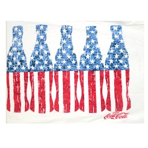 May include: White t-shirt with a design of Coca-Cola bottles in the style of the American flag. The bottles are blue with white stars, and the lower part has red and white stripes. The Coca-Cola logo is visible.