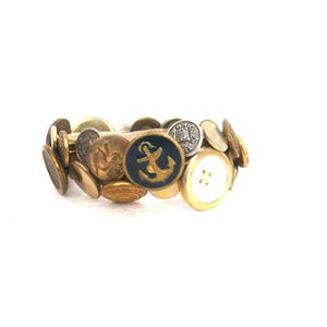 May include: A button bracelet featuring a variety of vintage buttons in gold, silver, and white. One navy blue button has a gold anchor design. The bracelet is on a white background.