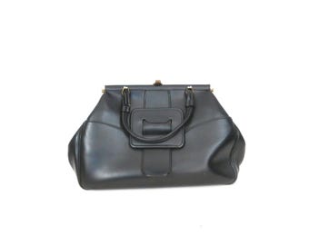 Puede incluir: Un bolso de mano negro con estructura rígida, que presenta dos asas redondeadas y un detalle de solapa rectangular central. El bolso tiene una silueta ancha y cónica con detalles de herrajes en tono dorado en el cierre y las asas.