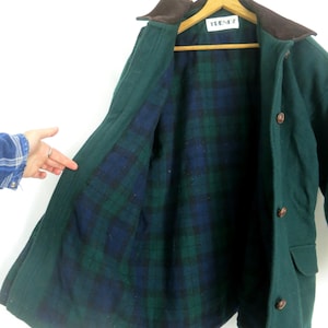 Puede incluir: Un abrigo de lana verde con cuello marr&oacute;n y forro a cuadros azul, verde y negro. El abrigo tiene botones marrones y una etiqueta que dice "TEENZ". Cuelga de una percha de madera.
