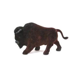 May include: A small, flocked figurine of a bison standing in a profile position. The animal features a dark brown body with a black head, neck, and horns. The surface has a soft, textured appearance, and the figure is depicted with a curved tail.