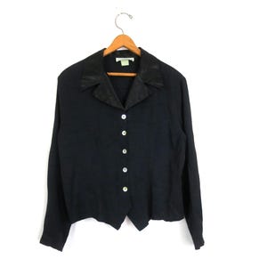 May include: Black long-sleeve button-down shirt with a contrasting dark collar. The shirt has a V-neck and is fastened with five white buttons. The garment is hanging on a wooden hanger against a white background.