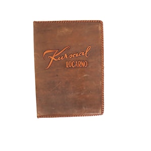 May include: A rectangular brown leather cover featuring a stitched border and the embossed text Kursaal LOCARNO on the front. The surface has a smooth, matte finish with natural variations in the brown tone across the entire front panel.