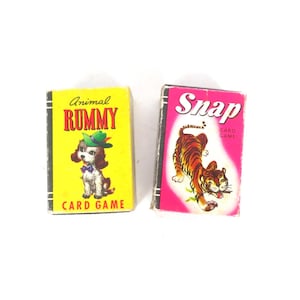 Può includere: Due scatole rettangolari di giochi di carte. La scatola gialla presenta un cucciolo con un cappello verde e il testo Animal RUMMY CARD GAME. La scatola rosa presenta un'illustrazione di una tigre e il testo Snap CARD GAME. Entrambe hanno dorsi neri.