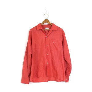May include: A long-sleeved button-down shirt in a solid coral red color. The shirt features a classic collar, a single chest pocket on the left side, and light-colored buttons running down the front placket and on the sleeve cuffs.