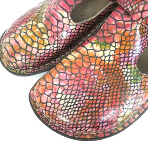May include: Close-up of a pair of colorful Mary Jane style shoes. The shoes feature a vibrant snakeskin pattern in shades of pink, orange, yellow, and black. The shoes have a strap across the top and a rounded toe.
