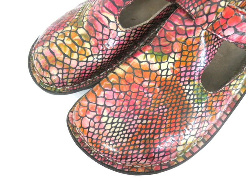 May include: Close-up of a pair of colorful Mary Jane style shoes. The shoes feature a vibrant snakeskin pattern in shades of pink, orange, yellow, and black. The shoes have a strap across the top and a rounded toe.