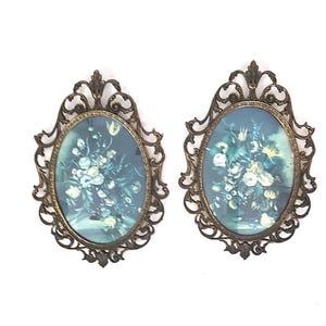 May include: A pair of oval wall frames featuring ornate, dark metallic scrollwork borders. Each frame contains a print of a floral bouquet with white and yellow flowers set against a dark, moody blue-green background, encased in glass.